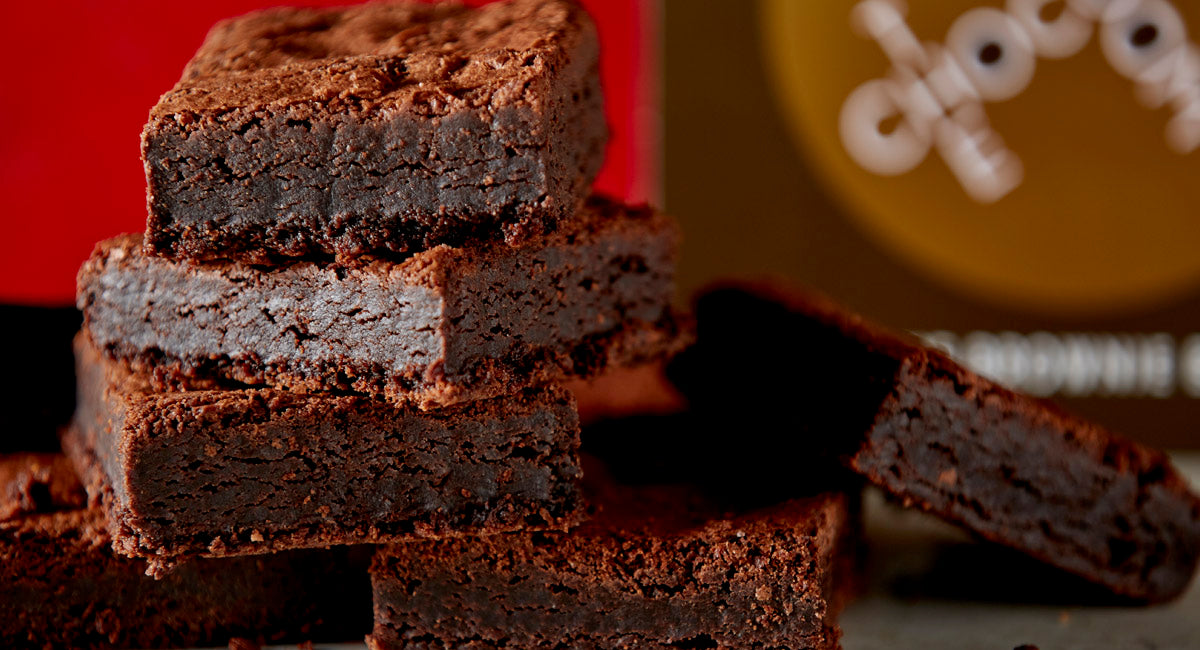 Close up of six stacked mouthwatering brownies in front of a stylish 12 piece brand box of brownies