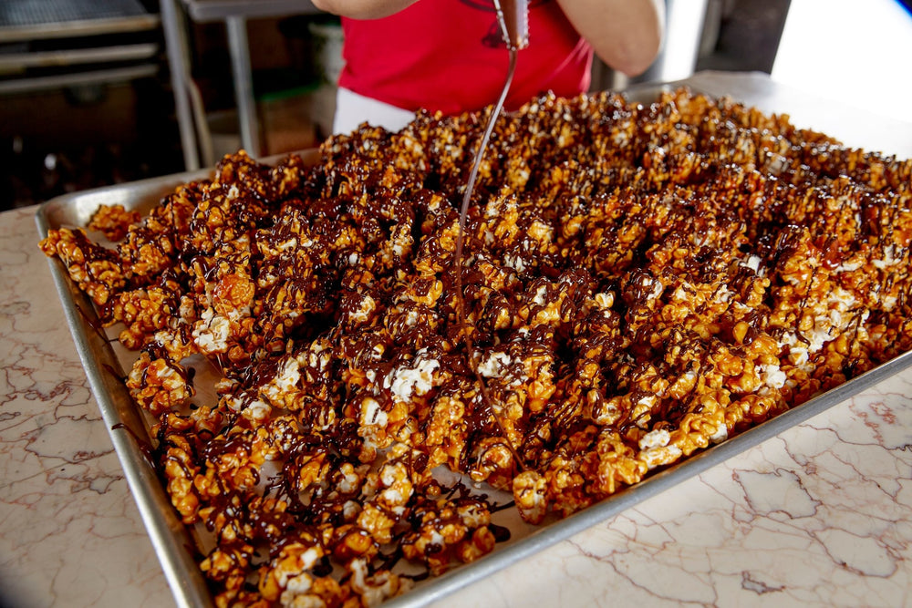 
                  
                    Chef piping chocolate over a sheet tray loaded with chocolate caramel popcorn. Sheet tray is on a marble counter
                  
                