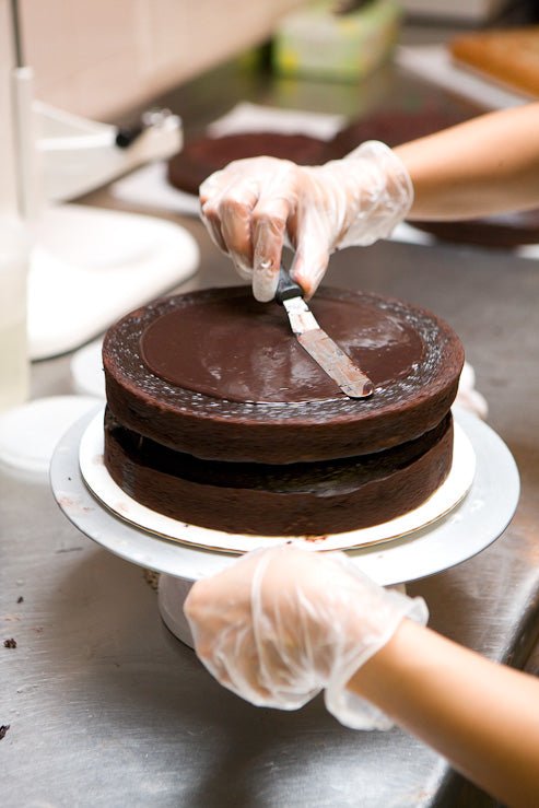 
                  
                    Chef staking the layers of a 6 inch layer on a frosting pedestal spreading blackout between the layers.
                  
                