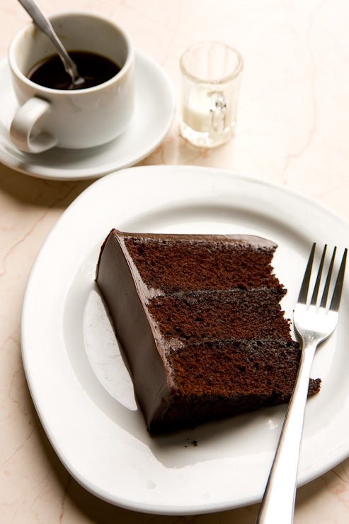 A slice of rich and moist chocolate layer cake on a plate with a fork next to mug of coffee