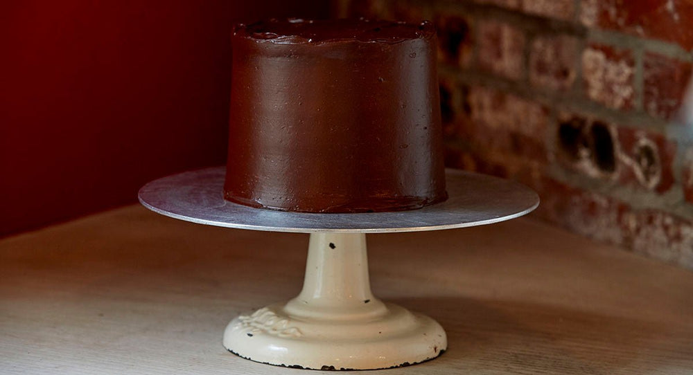 Perfectly frosted 6 inch chocolate layer cake on a frosting pedestal in front of a corner where a red and brick wall meet