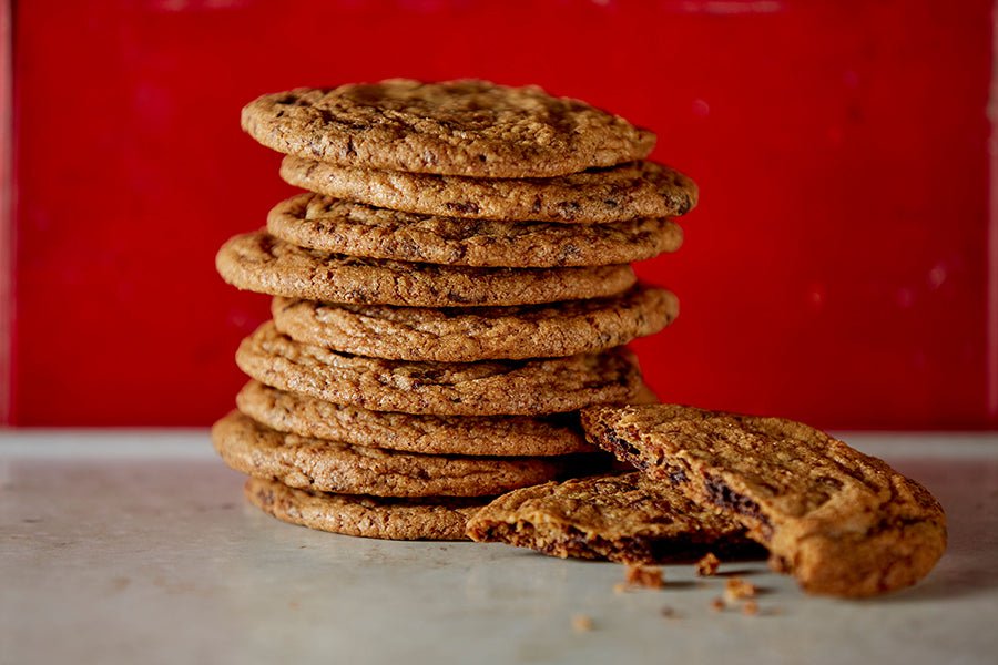 A stack of 9 crispy chocolate chip cookies with one cookie broken in half in front crumbs, on the counter, red background