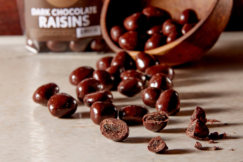 A tilted wooden bowl of dark chocolate raisons with some spilled on the counter. Some are cut open showing the raisons inside