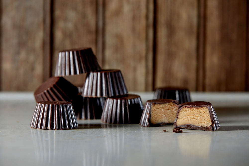 A pile of dark chocolate peanut butter cups with 1 cut open. They are on a vintage tabletop in front of an aged panned wall