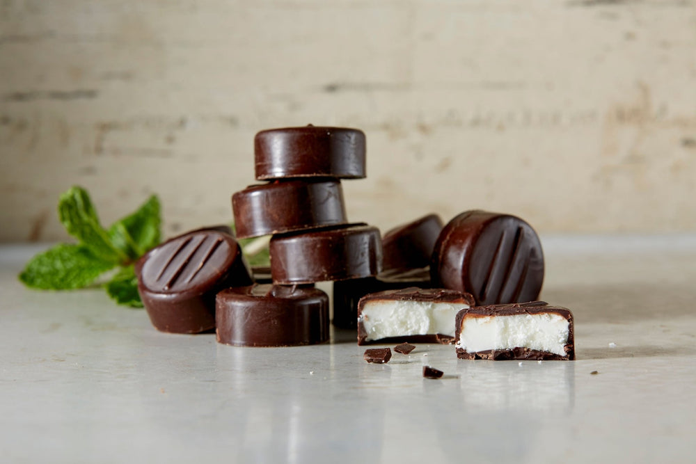 
                  
                    A stack of our dark chocolate peppermint patties. 1 is cut in half. Chocolate crumbs and fresh mint on the table
                  
                