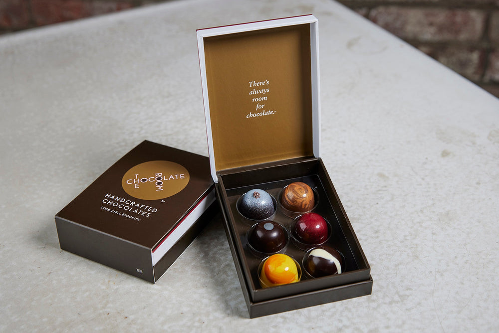 Open box of 6 handcrafted, colorful bonbons alongside a branded box - inside says There is always room of chocolate