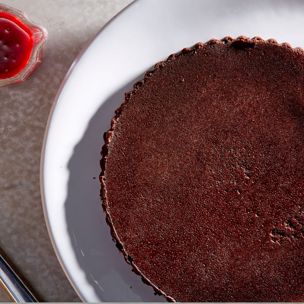 Overhead whole gluten free Flourless Chocolate Cake on a white plate next to a raspberry sauce boat and a silver cake slicer