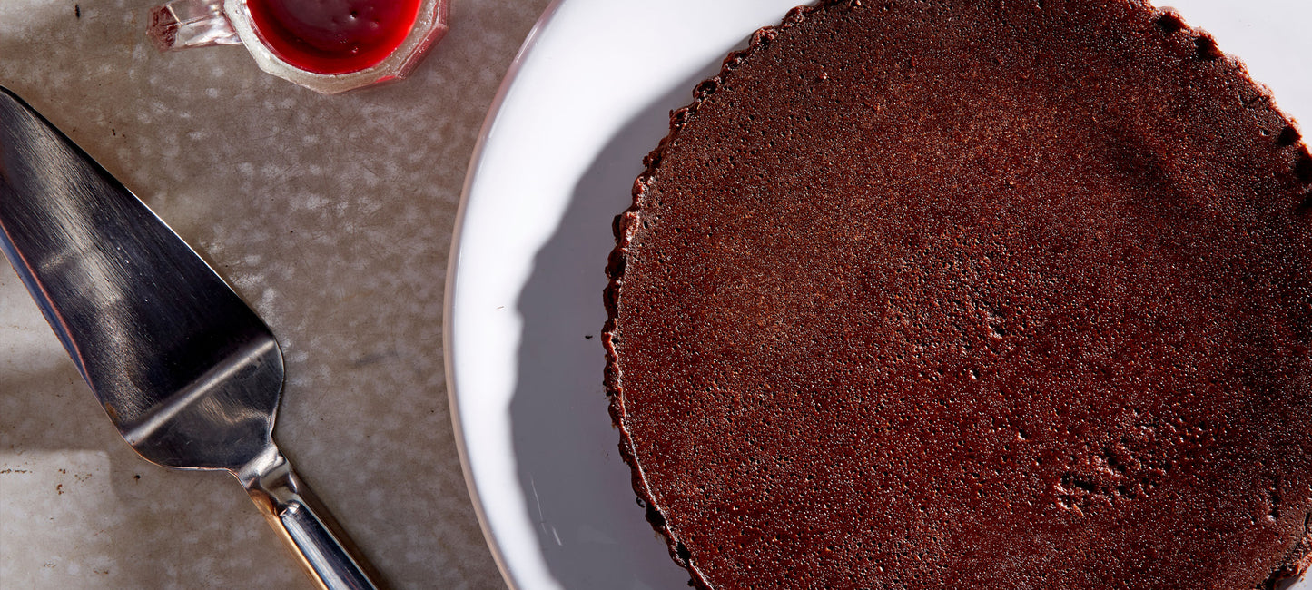 Overhead whole gluten free Flourless Chocolate Cake on a white plate next to a raspberry sauce boat and a silver cake slicer