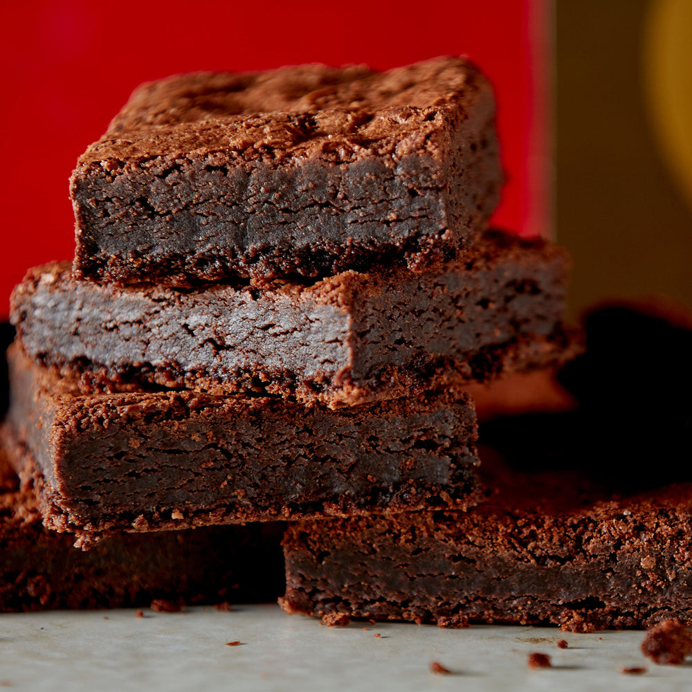 Close up of six stacked mouthwatering brownies in front of a stylish 12 piece brand box of brownies