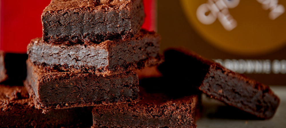 Close up of six stacked mouthwatering brownies in front of a stylish 12 piece brand box of brownies