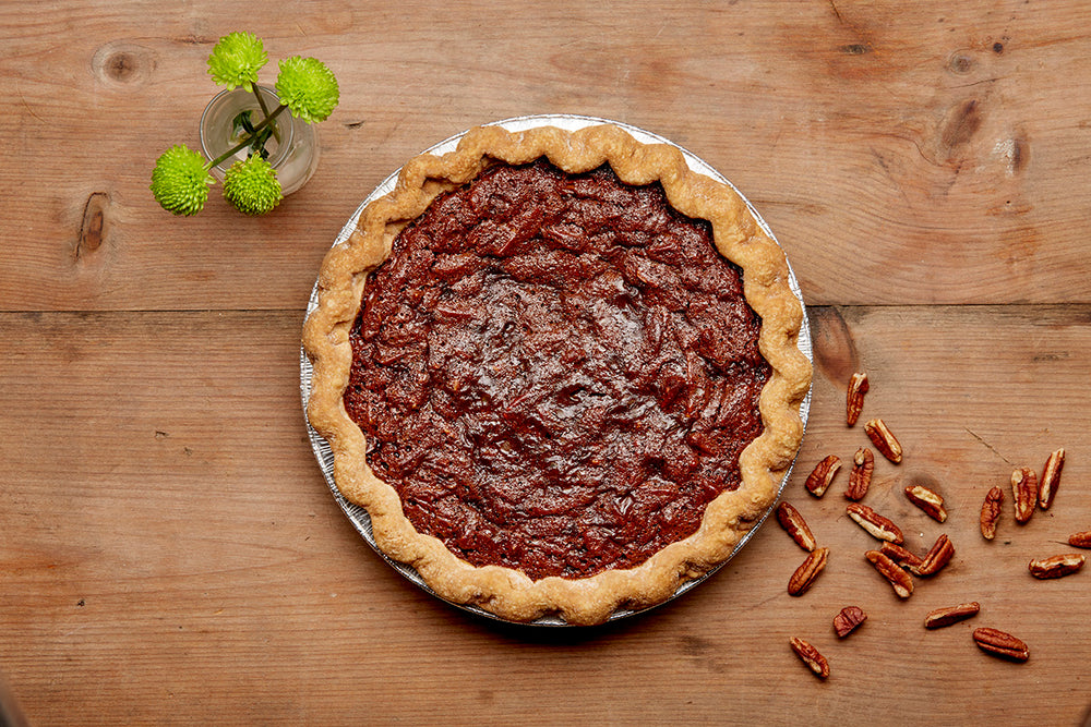 Overhead shot of our chocolate pecan pie on a wooden tabletop with a scattered pecans on it and small vase with green flowers